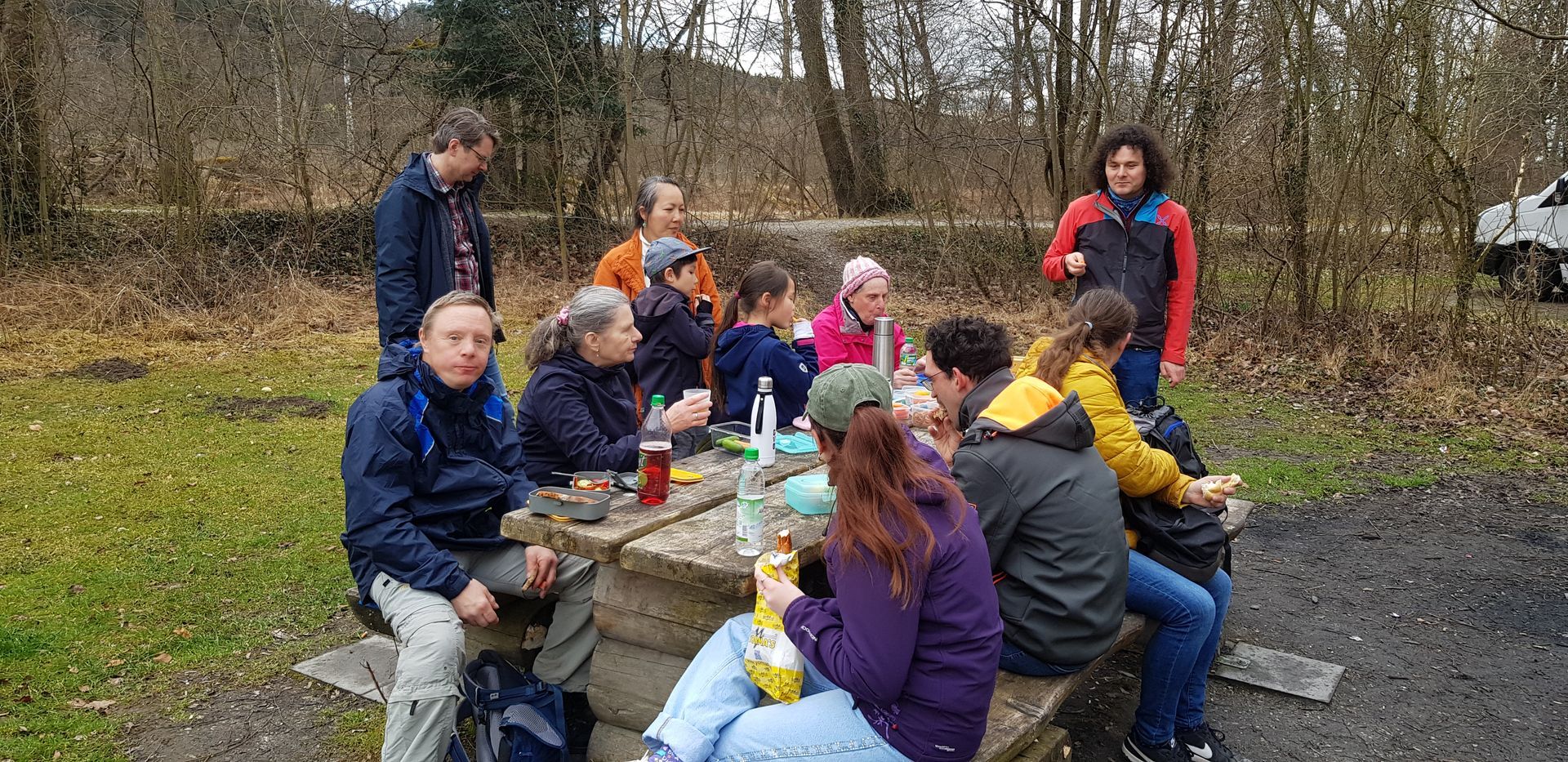 Gruppe von Kindern und Erwachsenen sitzt und steht um einen Picknicktisch im Wald, einige essen und trinken, andere unterhalten sich.