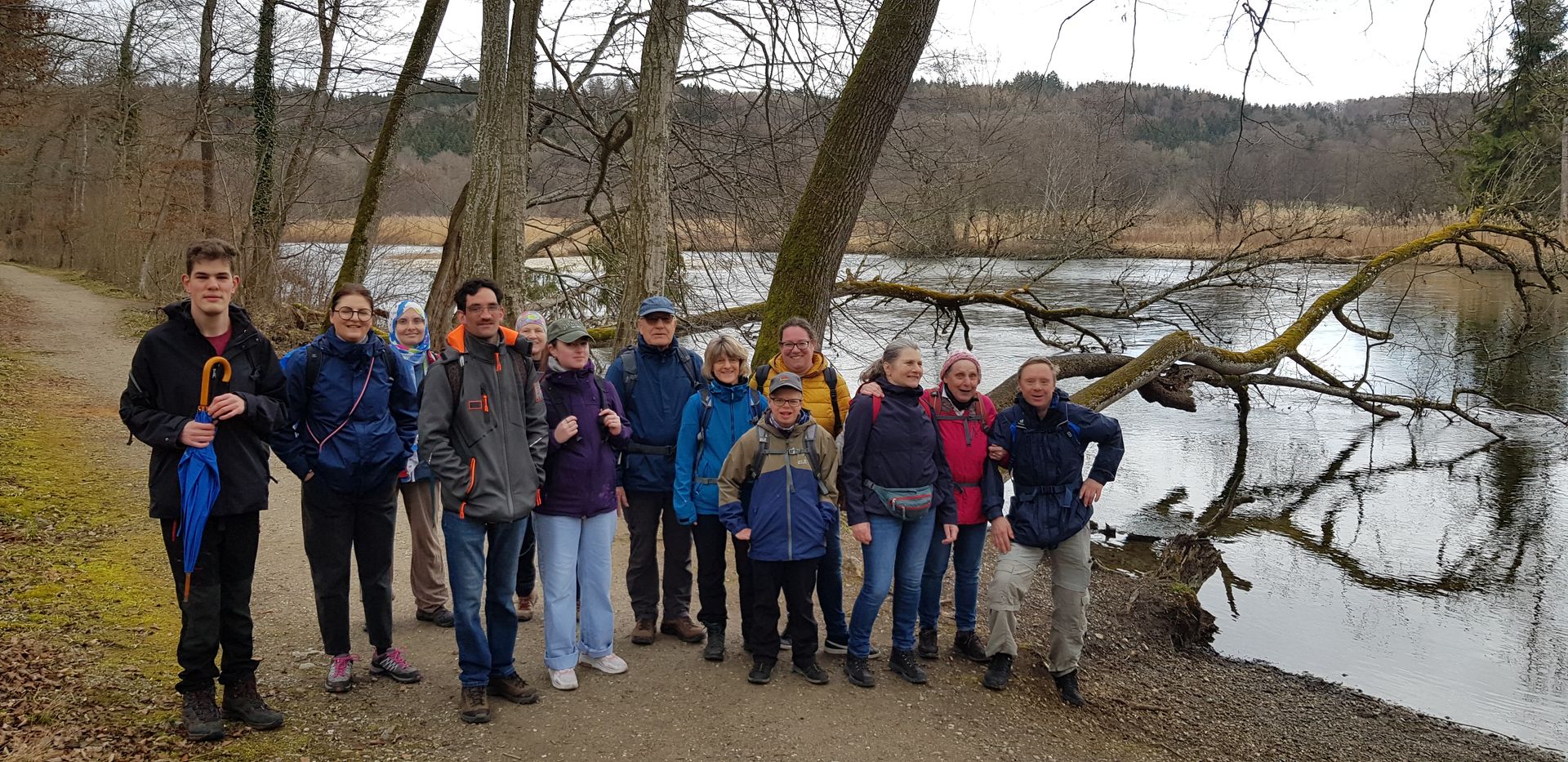 Gruppe von zwölf Personen mit Outdoor-Kleidung und Rucksäcken steht auf einem Waldweg neben einem See mit kahlen Bäumen im Hintergrund