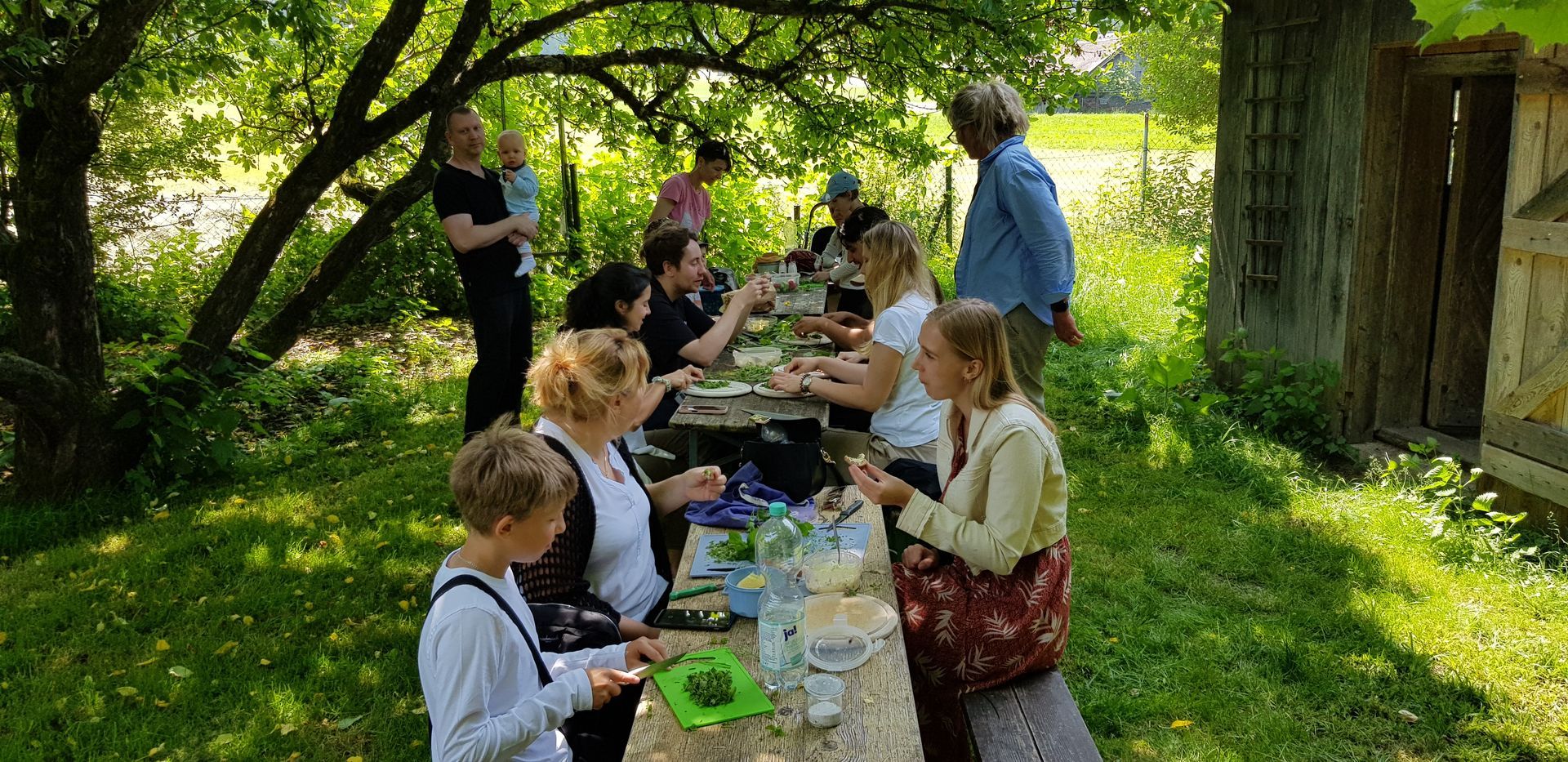 Mehrere Personen sitzen an einem langen Holztisch im Garten und bereiten gemeinsam Essen zu, umgeben von Bäumen und Gras.