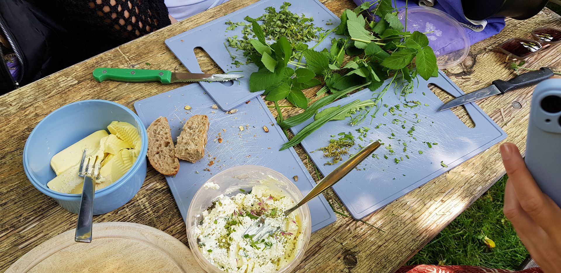 Frische Kräuter auf Schneidebrettern, Schüssel mit Kräuterquark, Butter in einer blauen Schüssel und zwei Brotscheiben auf einem Holztisch