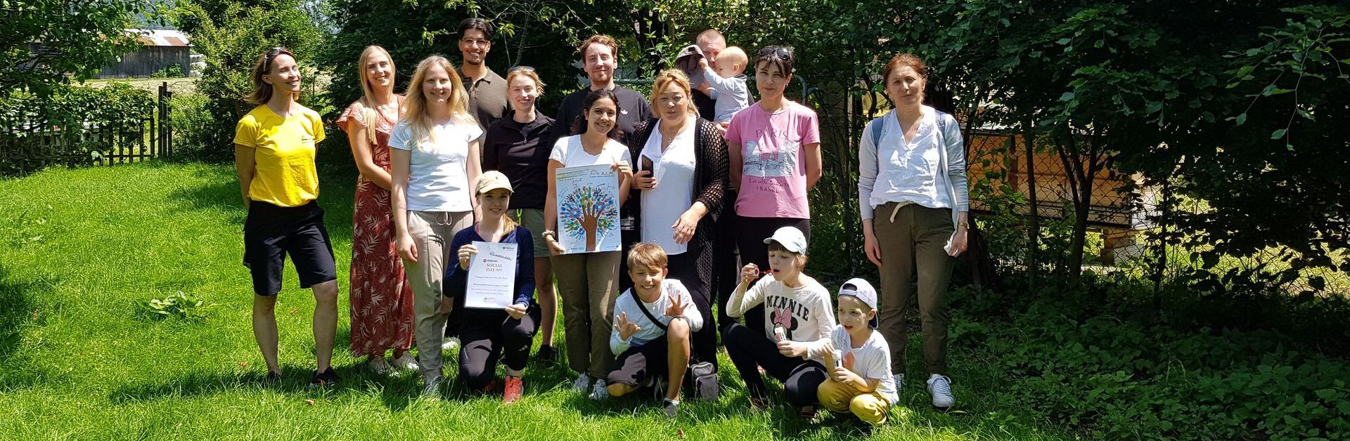 Gruppenfoto von 15 Personen auf einer Wiese mit Bäumen im Hintergrund, einige halten Urkunden und ein Bild mit einem Baum
