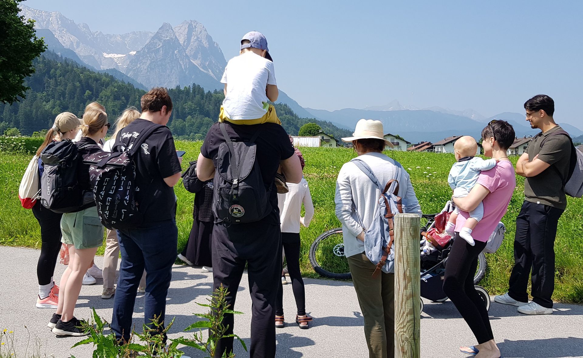 Gruppe von Menschen auf einem Wanderweg mit Bergen im Hintergrund, darunter ein Kind auf den Schultern eines Erwachsenen und eine Frau, die ein Kleinkind trägt.