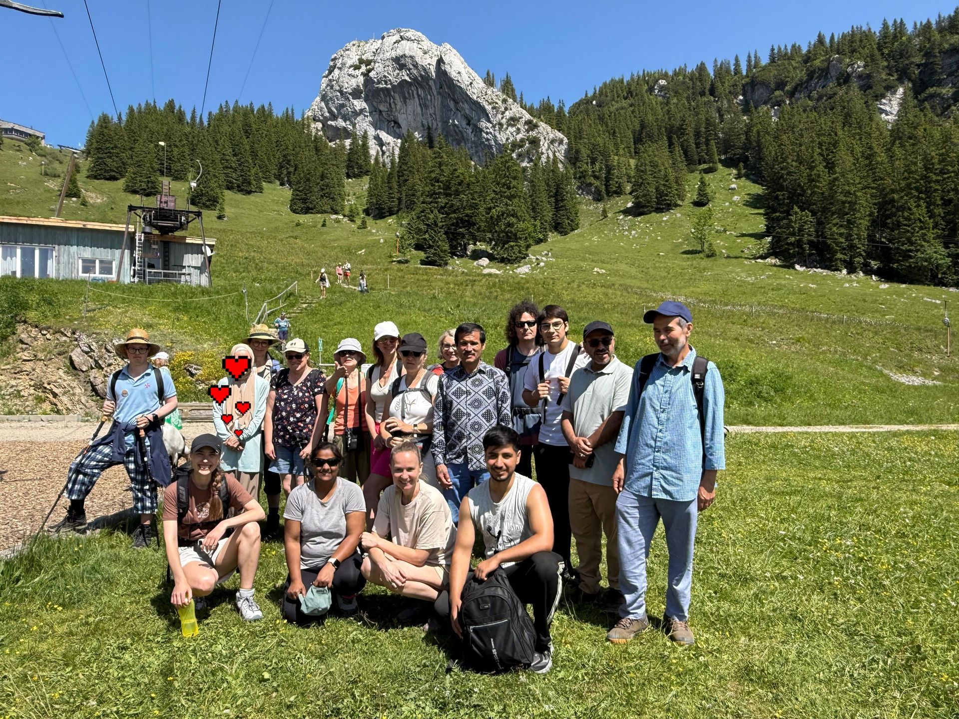 Gruppe von 15 Personen auf einer grünen Wiese vor einem bewaldeten Berg mit Felsgipfel bei sonnigem Wetter