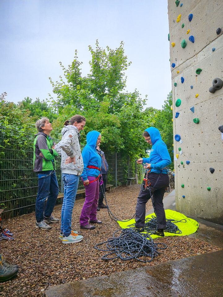 Es sind fünf Menschen auf dem Bild zu sehen. Sie bereiten sich fürs Klettern vor. Eine Person ist bereits am Sicherungsgerät eingebunden.
