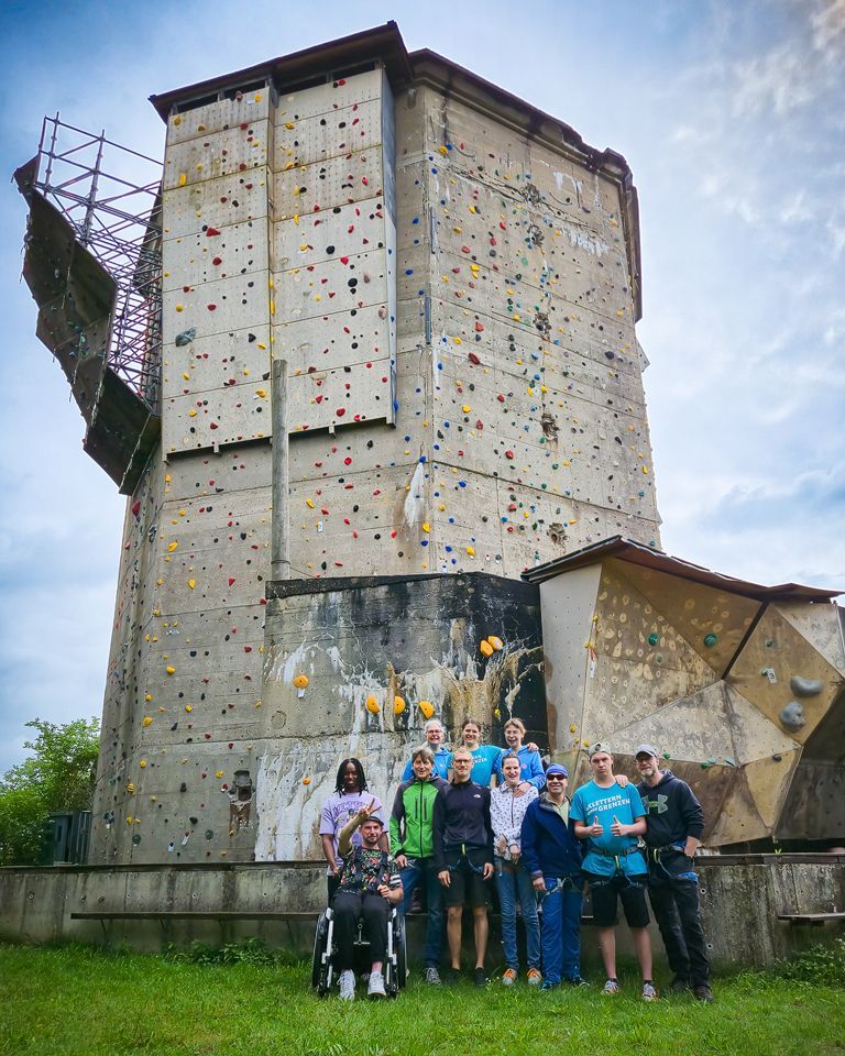 Es ist eine Gruppe von Menschen im Vordergrund zu sehen. Sie stehen auf einer Wiese. Alle tragen einen Klettergurt. Eine Person sitzt im Rollstuhl. Im Hintergrund ist eine Außenkletteranlage zu sehen.