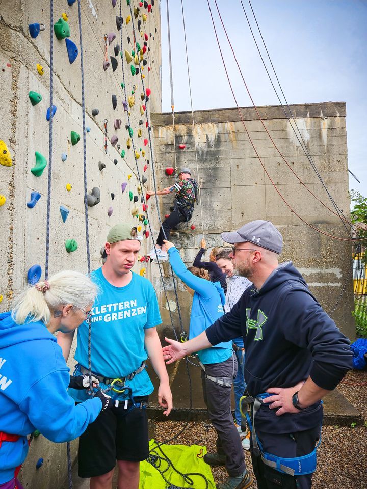 Es sind im Vordergrund drei Personen zu sehen. Eine Person seilt eine andere Person am Klettergurt an. Die Person rechts im Bild berührt den Unterarm der Person.