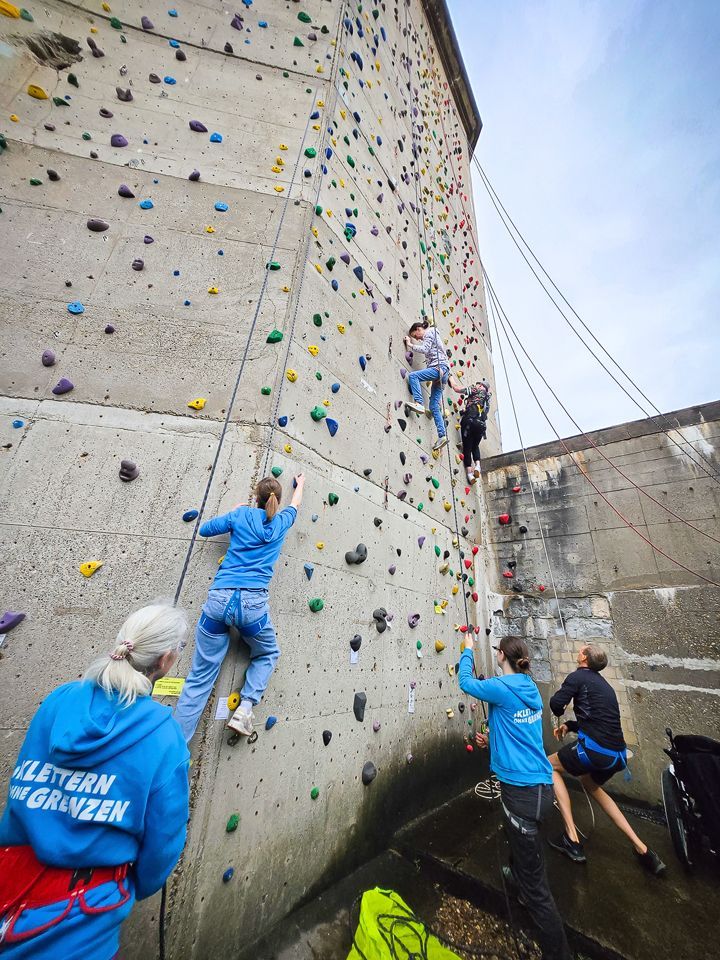Es ist die Außenkletteranlage zu sehen. Es sind drei Personen zu sehen, die drei weitere Kletternde an der Wand sichern.