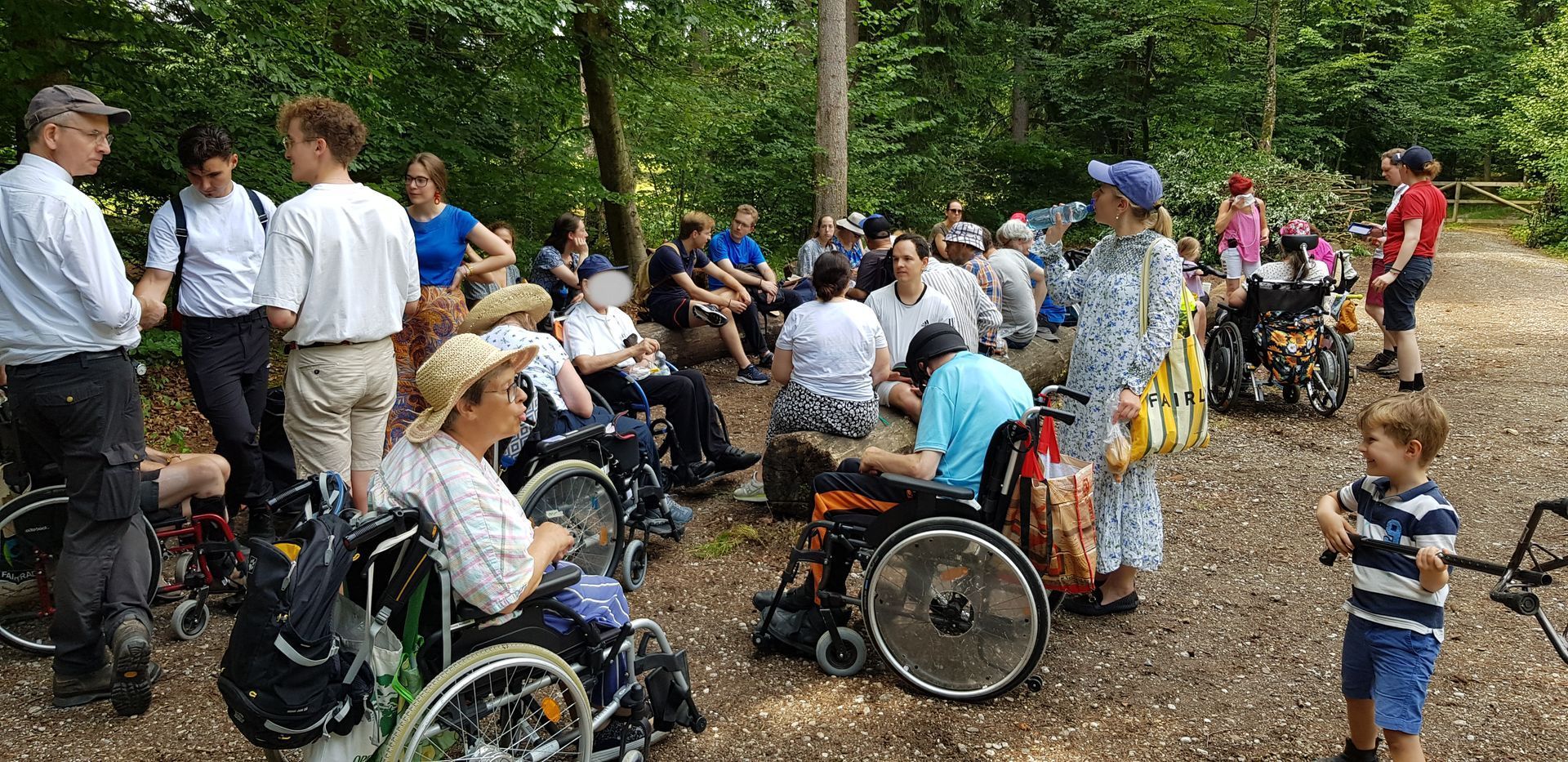 Gruppe von Menschen, darunter viele im Rollstuhl, versammelt auf einem Waldweg, einige stehen und andere sitzen, umgeben von Bäumen
