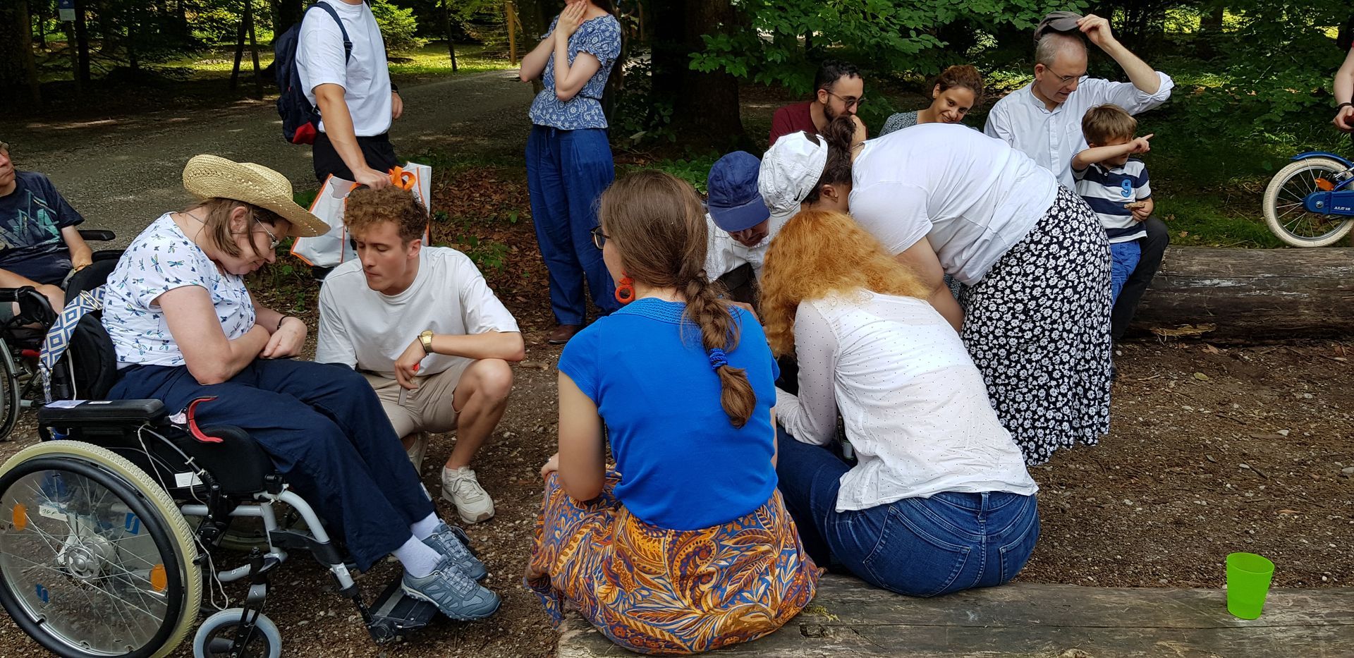 Frau im Rollstuhl mit Strohhut spricht mit junger Person in weißem T-Shirt, Sie sind umgeben von weiteren Menschen in Park mit Bäumen