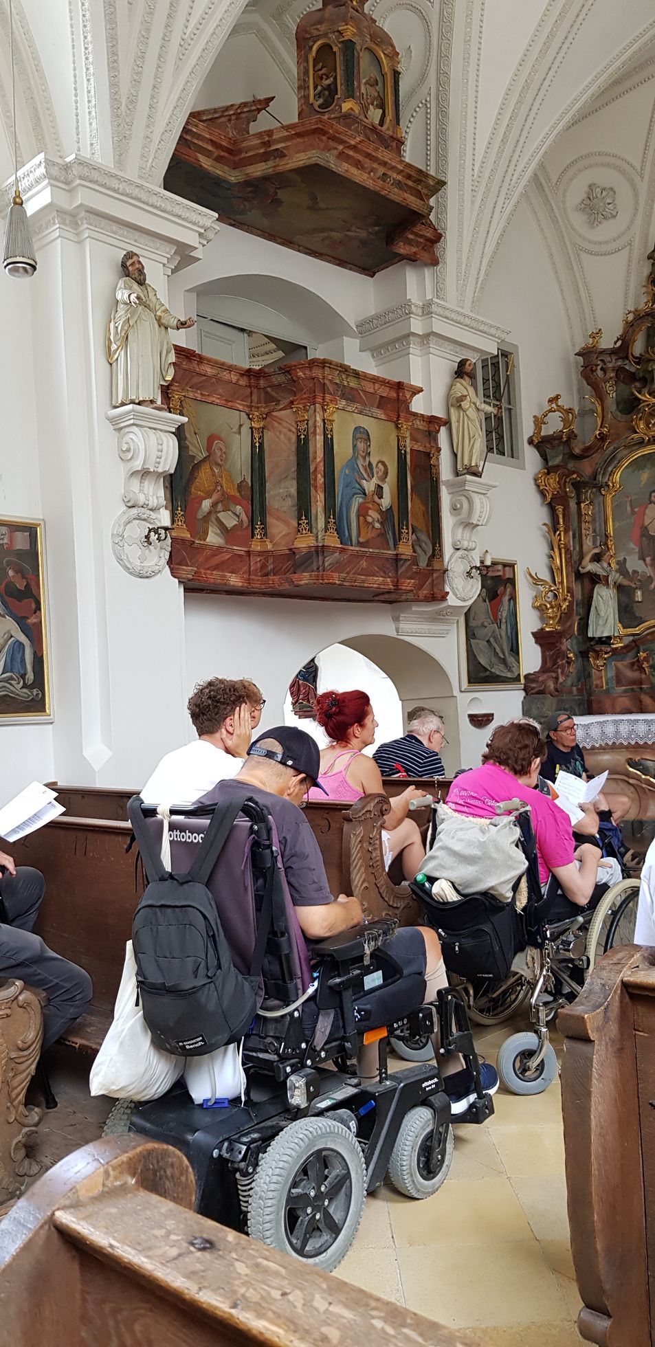Menschen in Rollstühlen und Sitzenden in einer Kirche mit weißen Wänden, barocken Altären und religiösen Gemälden