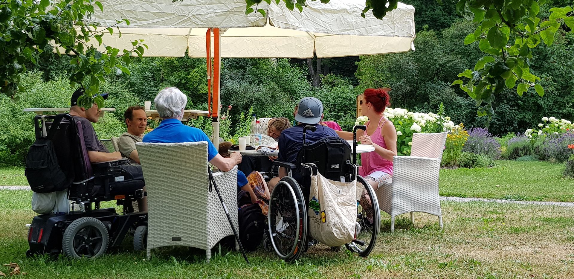 Gruppe von fünf Personen, darunter zwei im Rollstuhl, sitzt unter einem Sonnenschirm an einem Gartentisch im Grünen