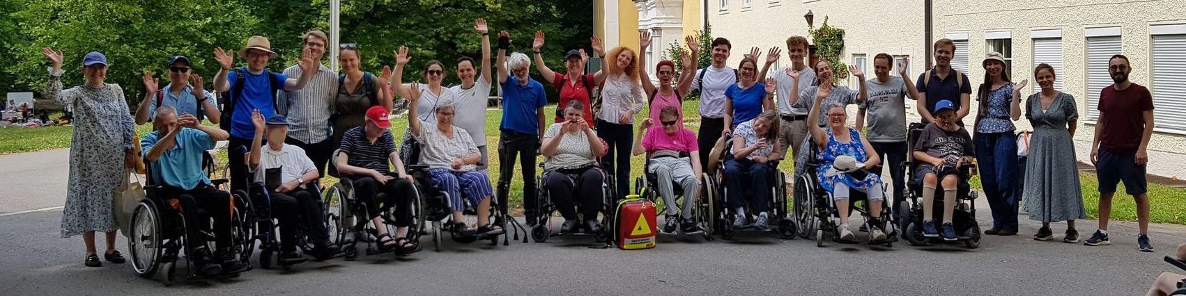 Gruppe von 29 Menschen, darunter 9 Rollstuhlfahrer, posieren zusammen vor einem gelben Gebäude im Freien. Sie winken in die Kamera.