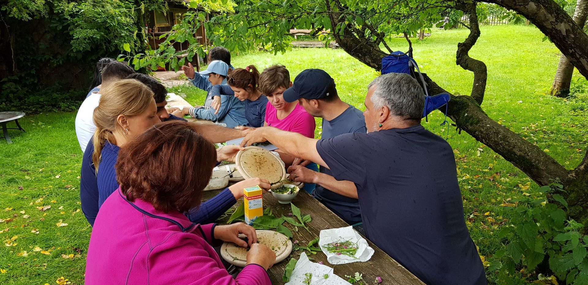 Gruppe von Menschen sitzt an einem langen Holztisch im Garten. Auf dem Tisch liegen Kräuter, Brot und Quark. Ein Frau schneidet Kräuter auf einem Brett. Eine andere Person gibt Kräuter in eine Schüssel mit Quark.