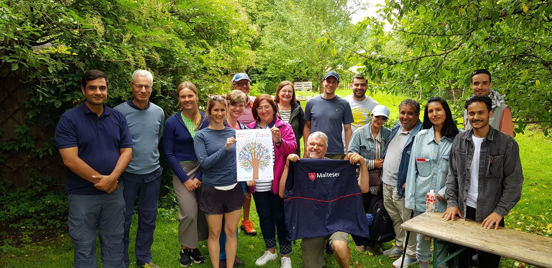 Gruppe von 15 Menschen steht und sitzt im Garten, eine Person hält ein buntes Baum-Plakat, eine weitere hält eine Jacke mit Malteser-Logo