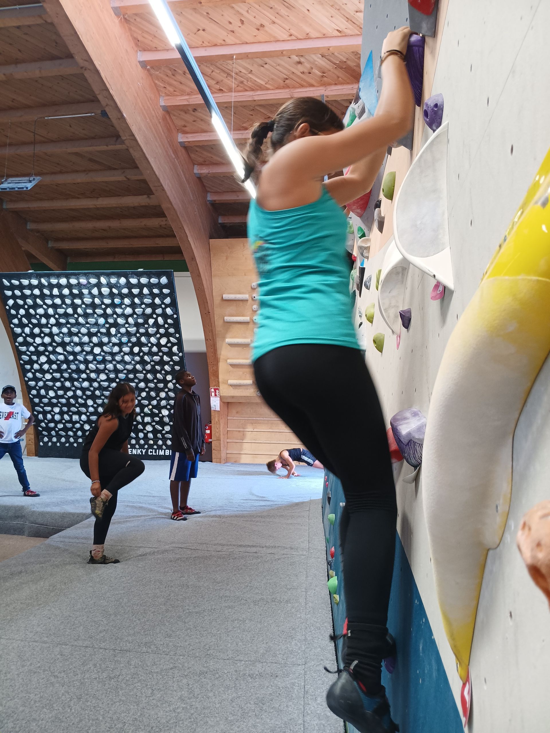 Frau in türkisfarbenem Tanktop und schwarzer Leggings klettert an einer Boulderwand in einer Halle