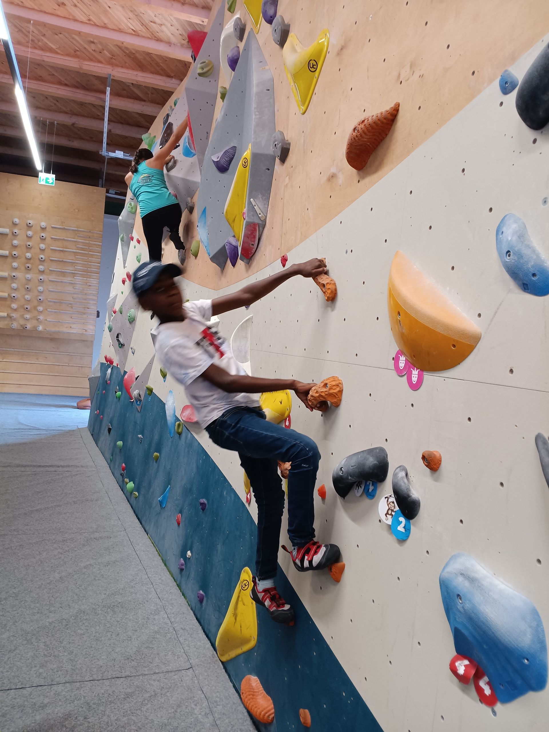 Im Vordergrund klettert ein Jugendlicher in weißem T-Shirt an einer bunten Kletterwand mit verschiedenen Griffen und Tritten in einer Halle. Im Hintergrund klettert eine Frau mit türkisfarbenem Tanktop an derselben Kletterwand.
