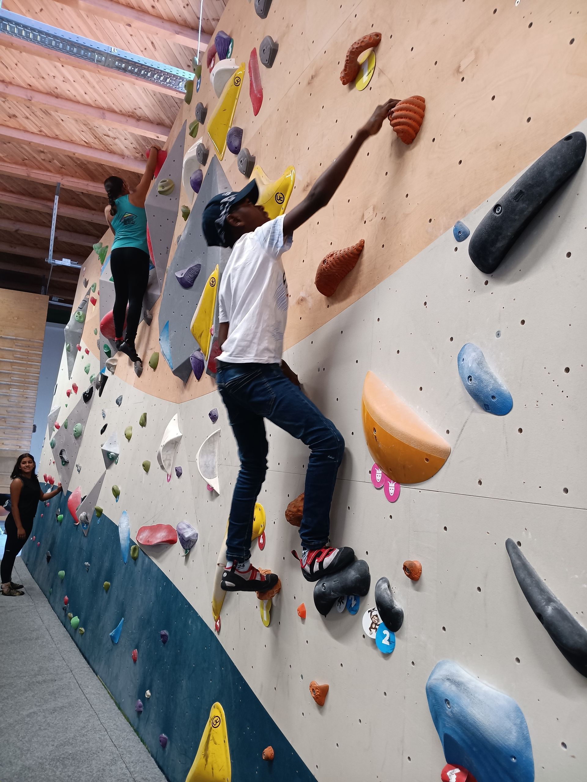 Zwei Personen klettern in Absprunghöhe an einer bunten Kletterwand mit verschiedenen Griffen und Tritten in einer Kletterhalle