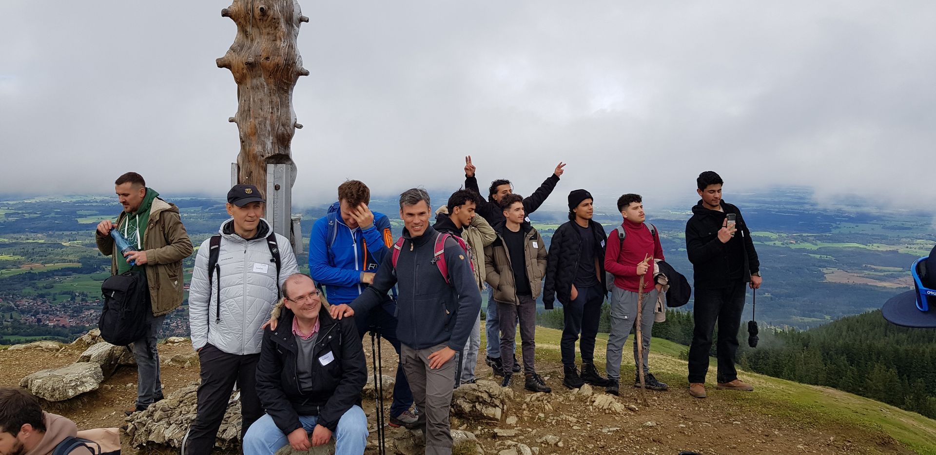 Gruppe von Personen in Outdoor-Kleidung auf felsigem Gipfel mit Baumstamm, im Hintergrund bewölkter Himmel und Landschaft mit Wäldern und Feldern.
