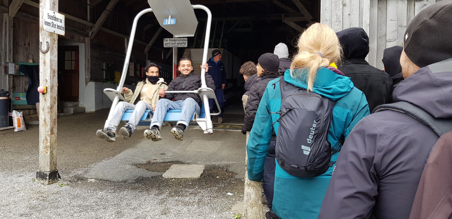 Menschen stehen in einer Schlange vor einer Seilbahnstation, zwei Personen sitzen auf einer Doppelsitz-Schwebebahn mit Metallrahmen.