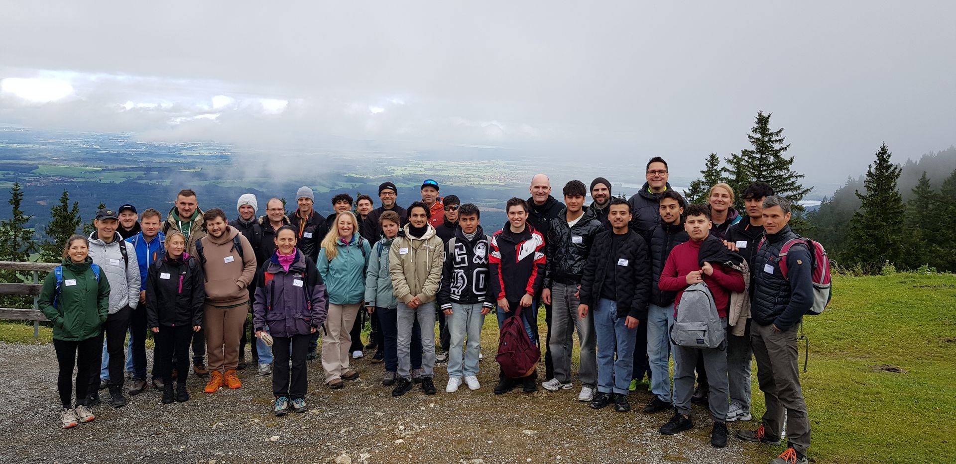 Große Gruppe von Menschen in Outdoor-Kleidung steht auf einem Weg mit Blick auf bewaldete Hügel und bewölkten Himmel im Hintergrund.