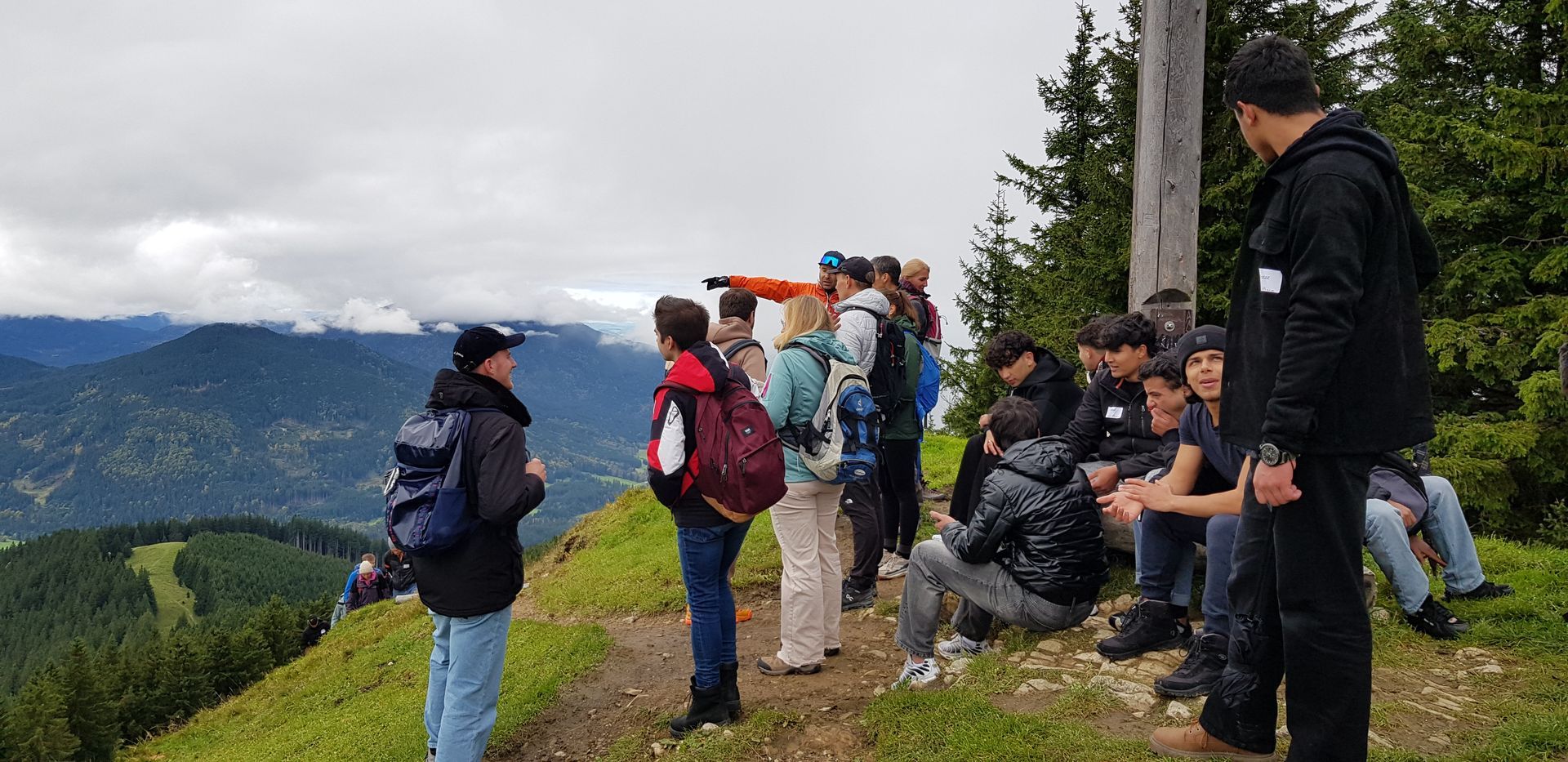Gruppe von Wanderern mit Rucksäcken auf einem Bergpfad, einige stehen, andere sitzen, im Hintergrund bewaldete Berge und bewölkter Himmel.
