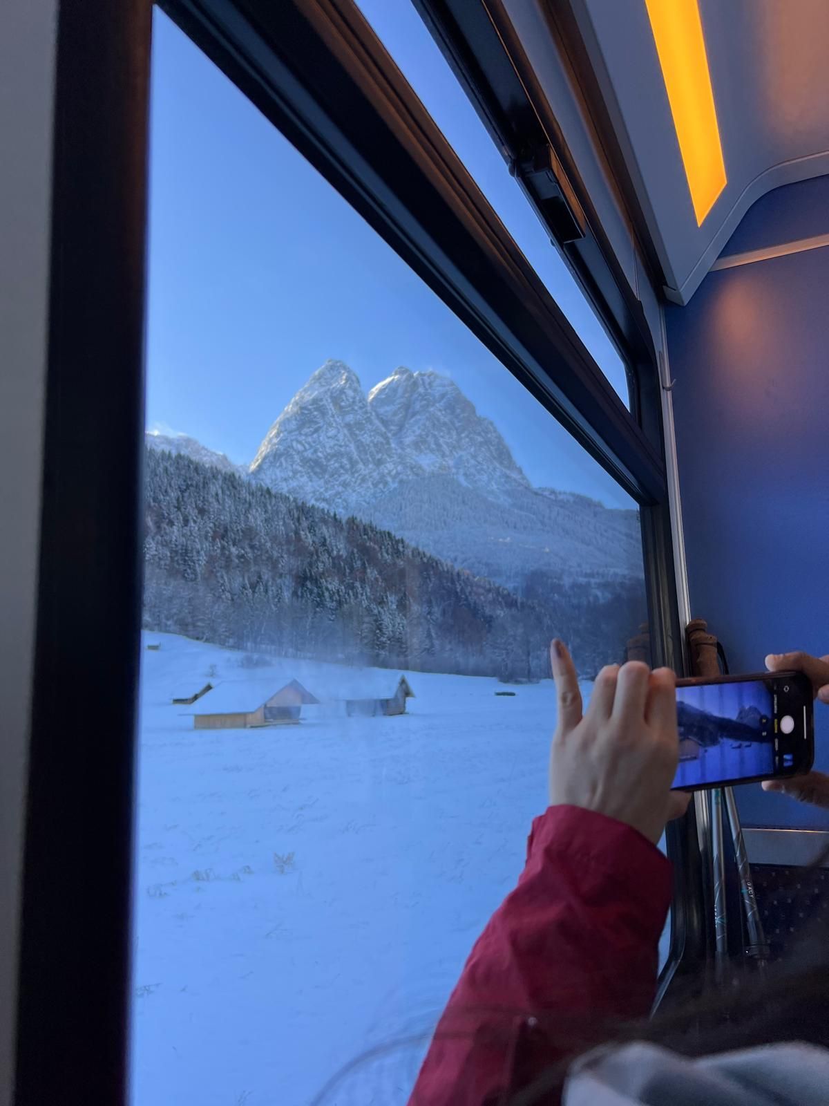 Blick aus einem Zugfenster auf schneebedeckte Hütten und Berge, eine Hand hält ein Smartphone zum Fotografieren.