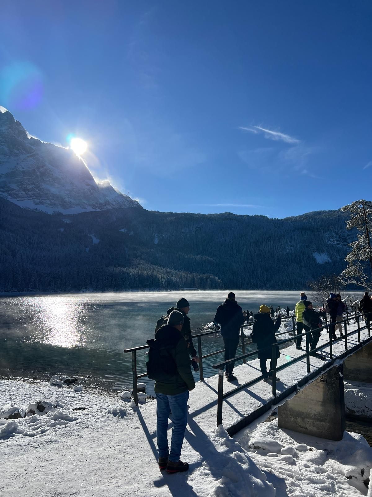 Menschen gehen auf einem schneebedeckten Steg neben einem zugefrorenen See, im Hintergrund Berge und Sonne am klaren Himmel.