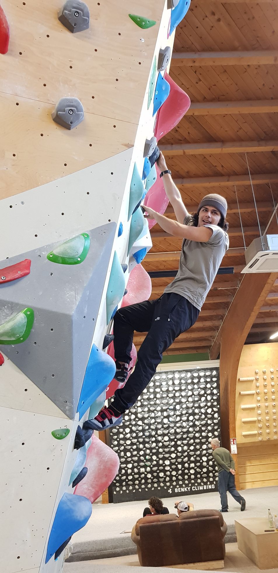 Person klettert an einer Kletterwand mit bunten Griffen in einer Halle mit Holzbalkendecke