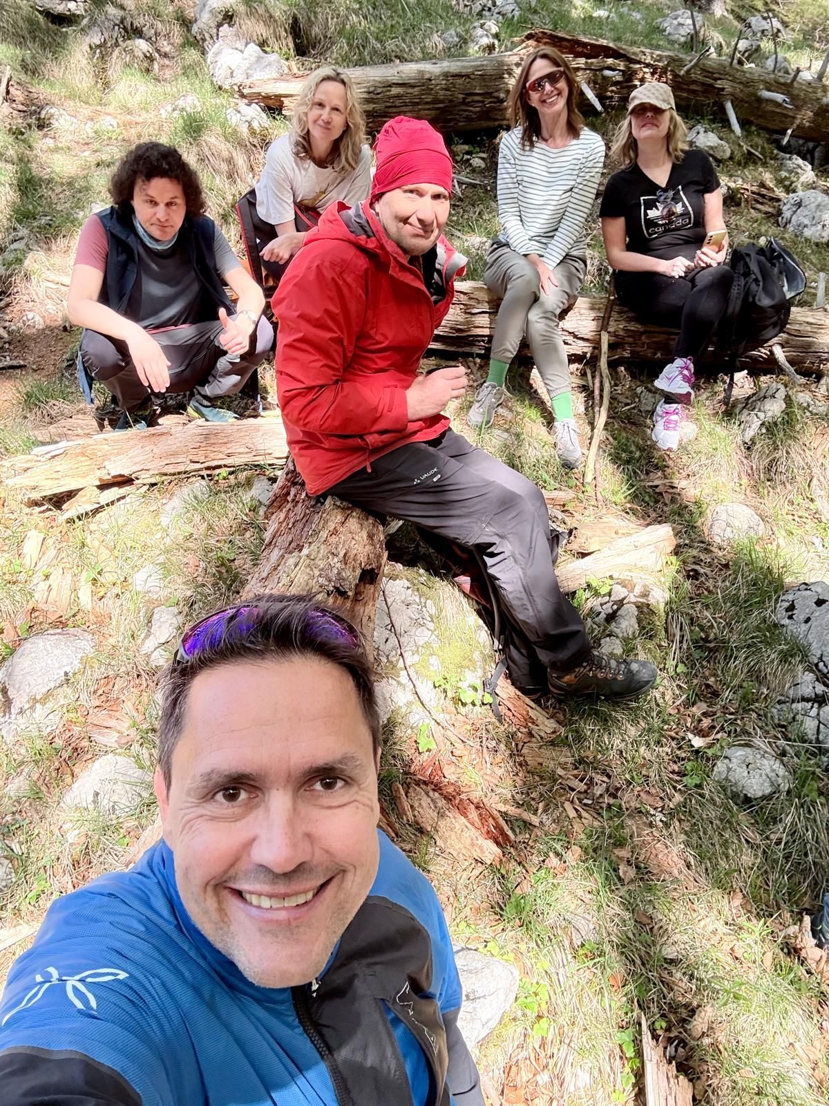 Fünf Personen sitzen auf umgestürzten Baumstämmen im lichten Wald. Eine weitere Person steht im Vordergrund und hält die Kamera für ein Gruppenfoto. Sonnenlicht fällt durch die Bäume und erzeugt helle Flecken auf dem Waldboden, der mit Gras, Steinen und Ästen bedeckt ist. Die Personen tragen Outdoor-Kleidung in verschiedenen Farben, darunter eine rote Jacke und eine blaue Jacke. Im Hintergrund sind Baumstämme und Felsen zu sehen.