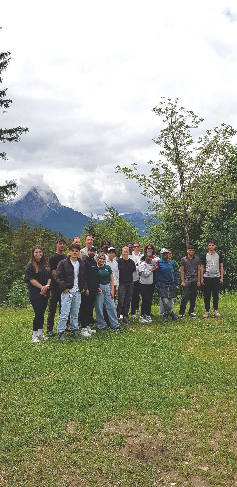 Eine Gruppe von Menschen steht auf einer Wiese vor einer Kulisse aus Bäumen und bewaldeten Bergen. Im Hintergrund sind wolkenverhangene Berge zu sehen. Die Personen stehen dicht beieinander, einige lächeln, andere blicken neutral in Richtung Kamera. Die Kleidung ist überwiegend sportlich und wetterfest, darunter Jacken, lange Hosen und Turnschuhe. Das Licht ist diffus, der Himmel ist größtenteils bewölkt.