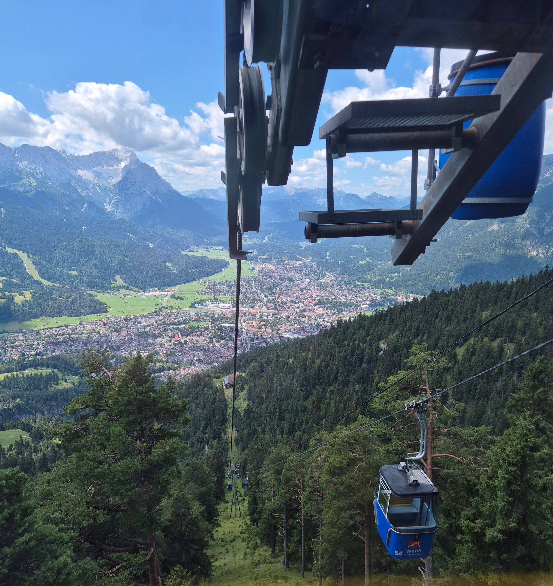 Blick aus einer Gondelbahn auf eine weitere blaue Gondel, darunter Wald und eine Stadt vor Bergkulisse unter blauem Himmel mit Wolken