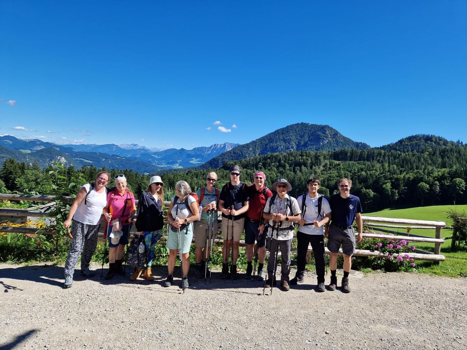 Eine Gruppe von zehn Personen steht auf einem Schotterweg in einer Berglandschaft. Im Hintergrund sind grüne Hügel und Berge sichtbar.