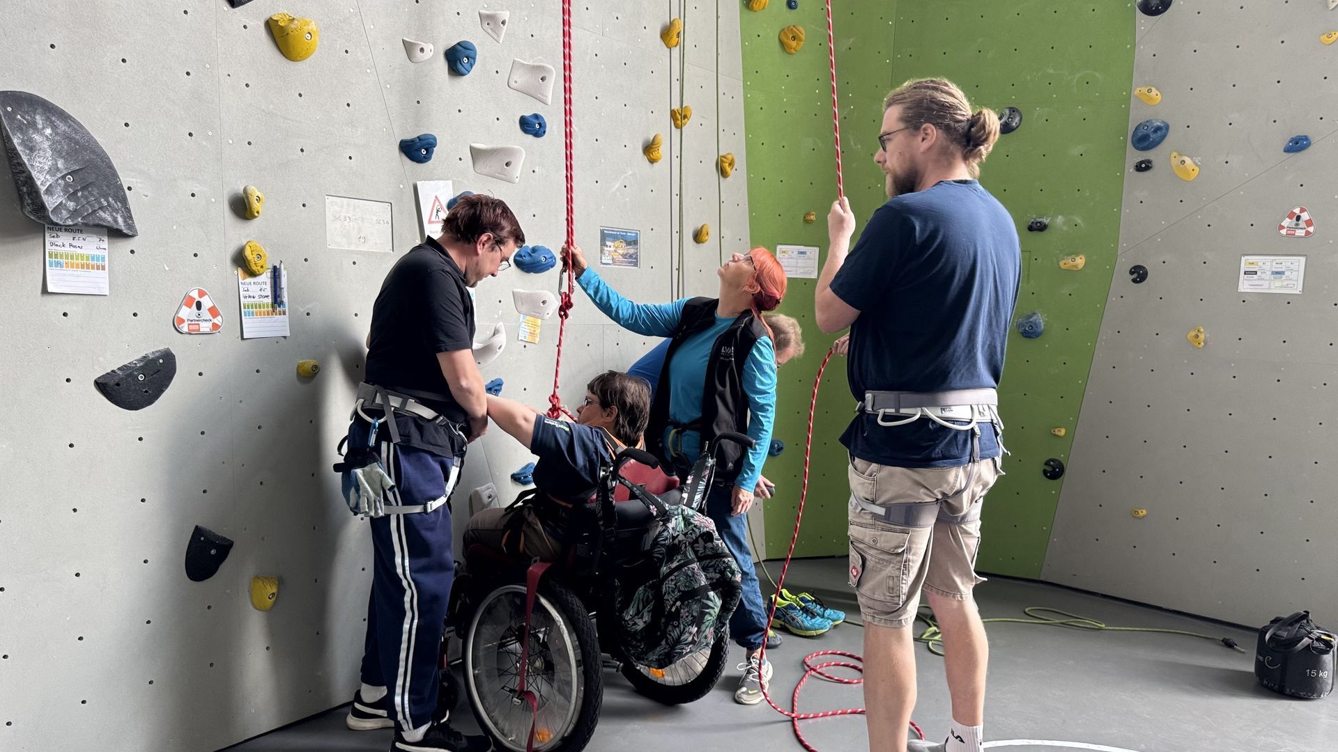 Eine Frau im Rollstuhl steht an der Kletterwand. Sie hat einen Klettergurt an. 3 Personen stehen zur Unterstützung bereit.
