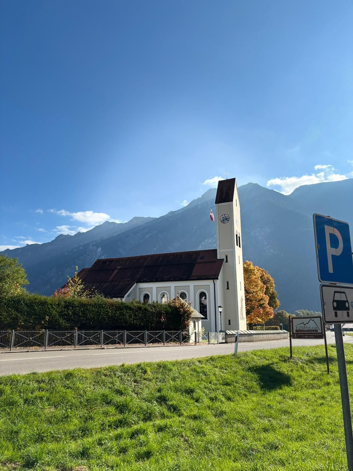 Kirche mit Turm und Uhr vor bewaldeten Bergen unter blauem Himmel, im Vordergrund eine grüne Wiese.