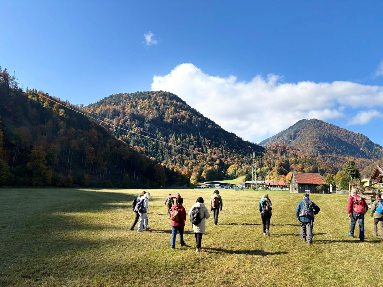 Eine Gruppe von Wanderern mit Rucksäcken auf einer Wiese vor bewaldeten, herbstlich gefärbten Bergen unter blauem Himmel mit Wolken.