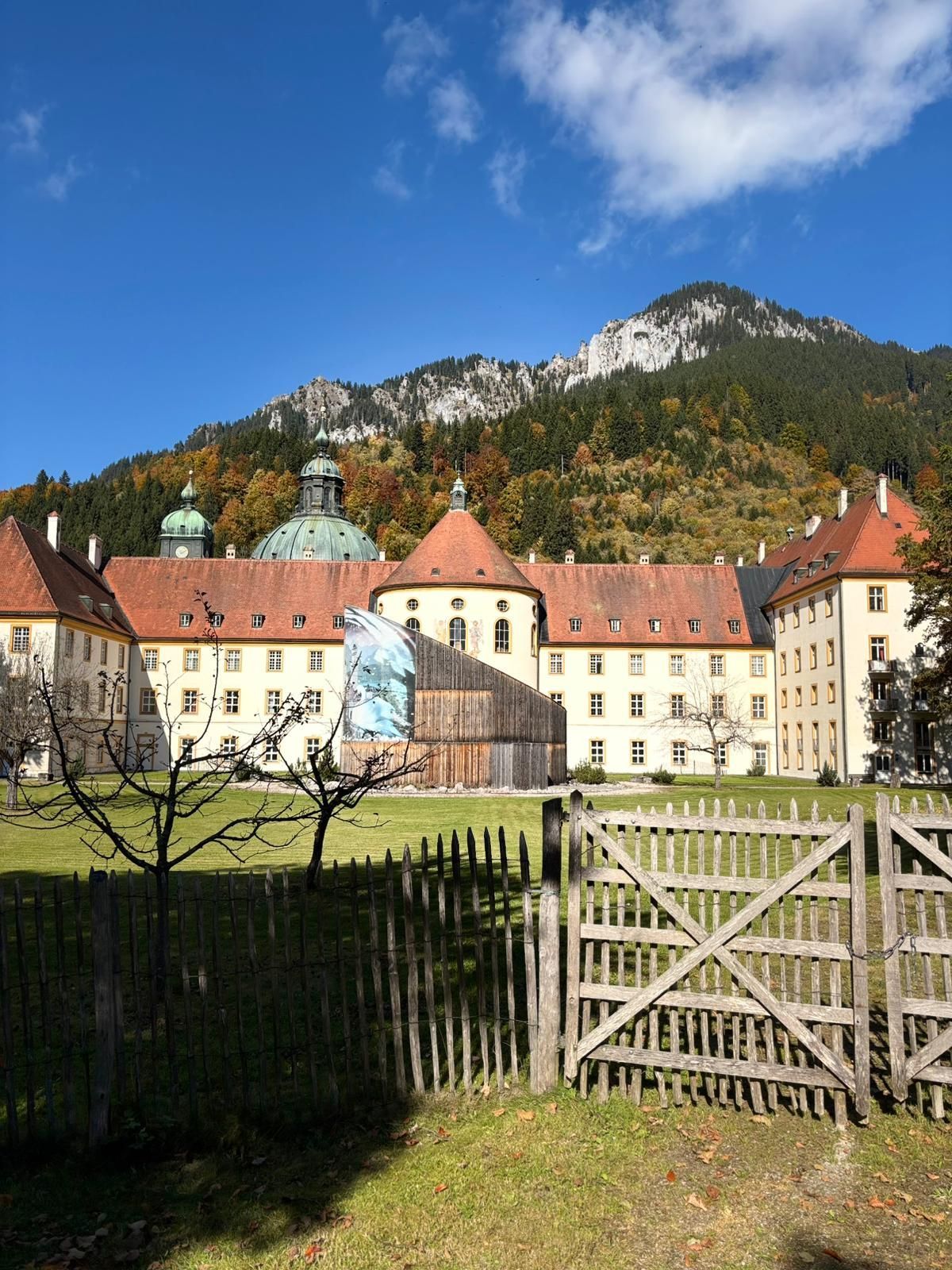 Historisches Kloster mit roten Dächern und grünen Kuppeln vor bewaldetem Berg unter blauem Himmel.