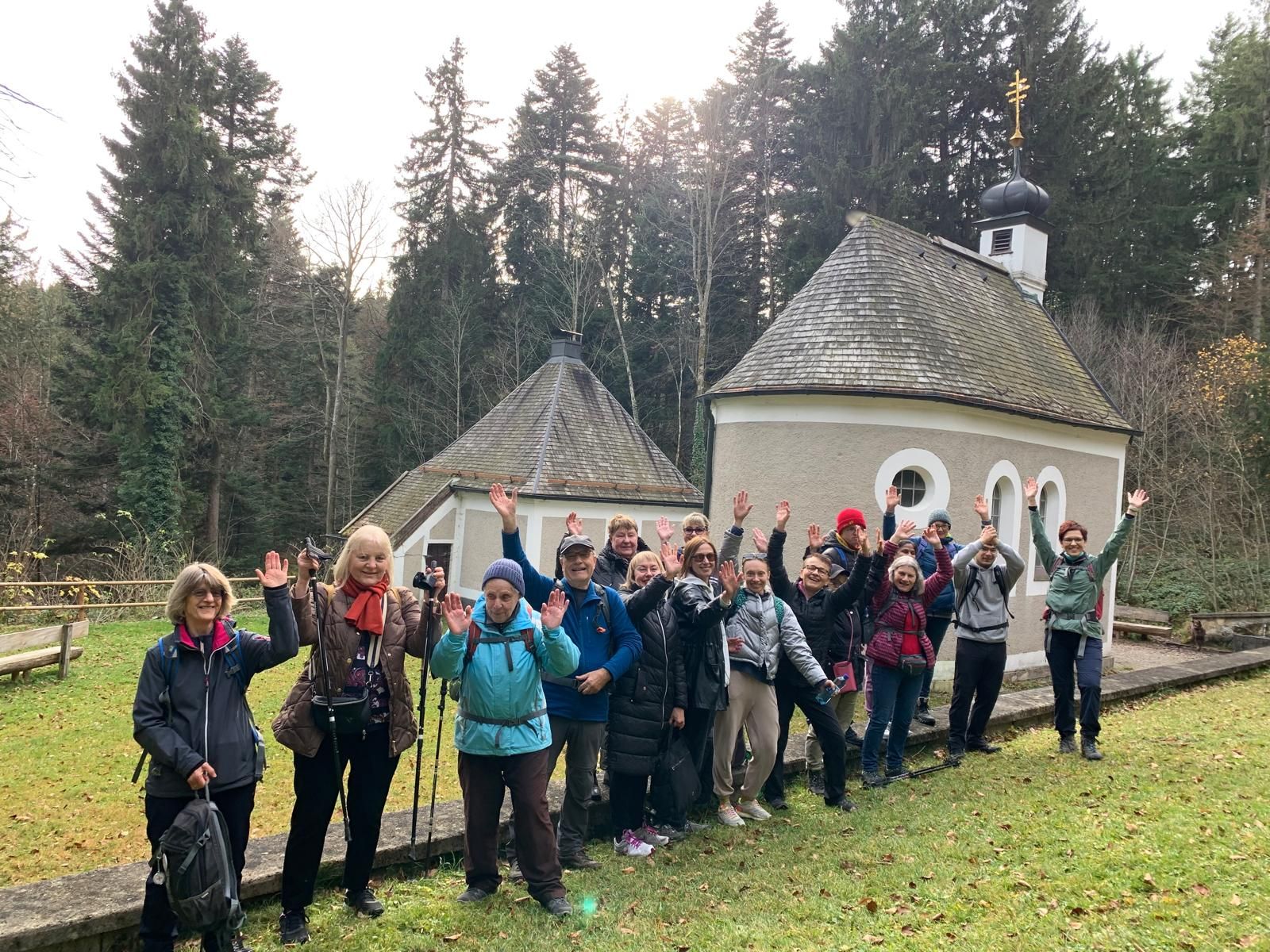 Gruppe von Menschen mit Wanderkleidung und Rucksäcken steht vor zwei kleinen Kapellen in einem Waldgebiet. Einige Wanderer heben die Arme.