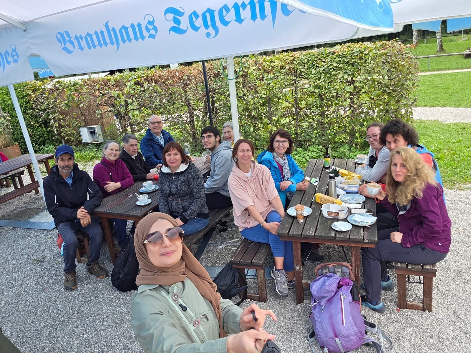 Eine Gruppe von Personen sitzt an einem Tisch im Biergarten. Auf dem Tisch stehen Teller, Gläser und eine Flasche. Die Personen tragen unterschiedliche Kleidung und sind in entspannter Haltung.