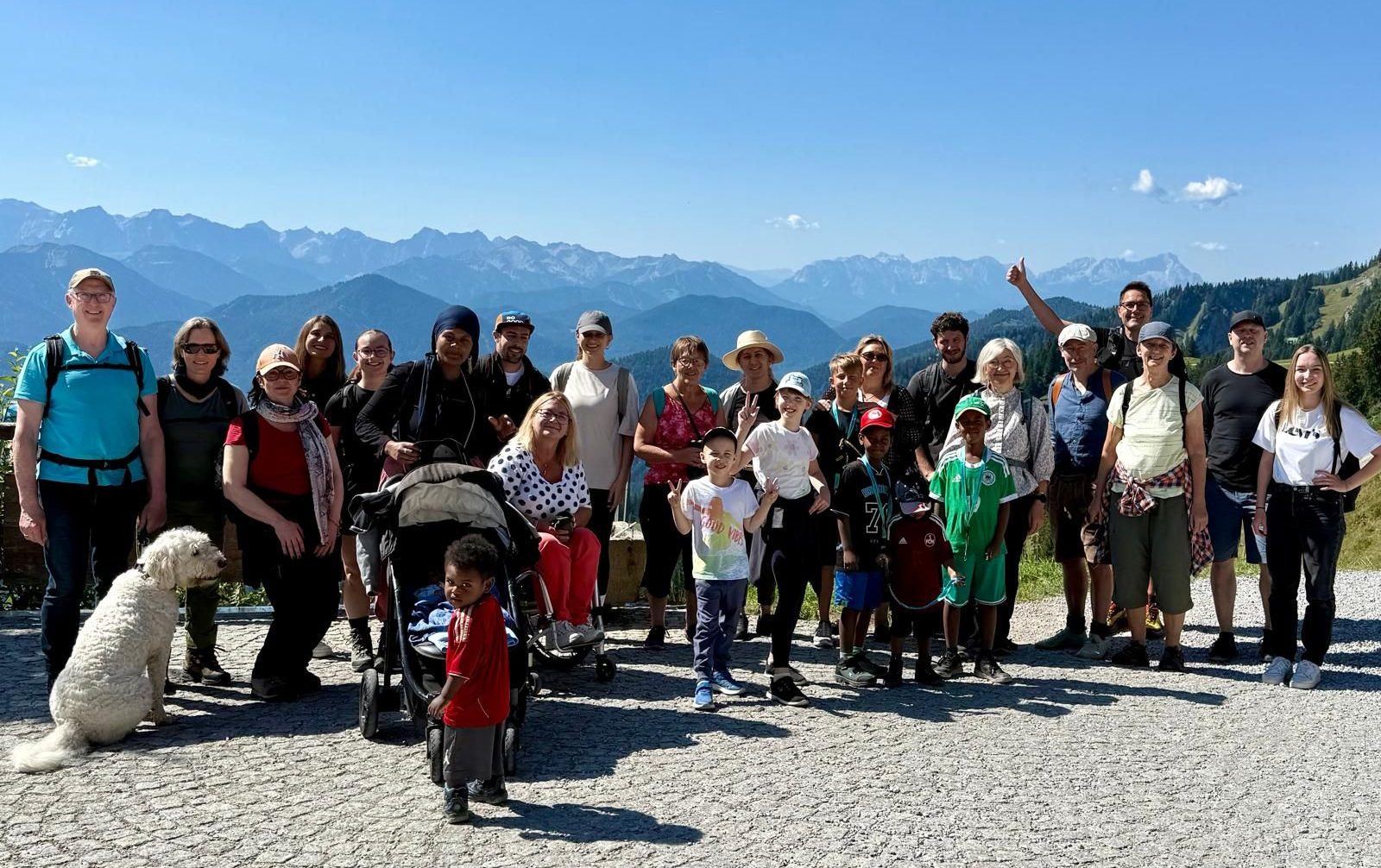 Gruppenfoto von 24 Personen, darunter Erwachsene und Kinder, in einer Berglandschaft mit schneebedeckten Gipfeln im Hintergrund. Ein Hund steht neben einem Kinderwagen.