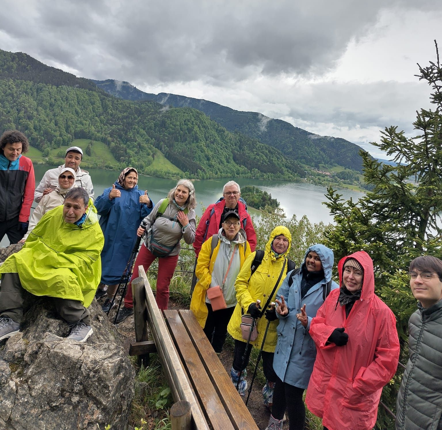 Gruppe von Menschen in bunter Outdoor-Bekleidung auf einem Felsen mit Blick auf bewaldete Berge und einen See unter bewölktem Himmel