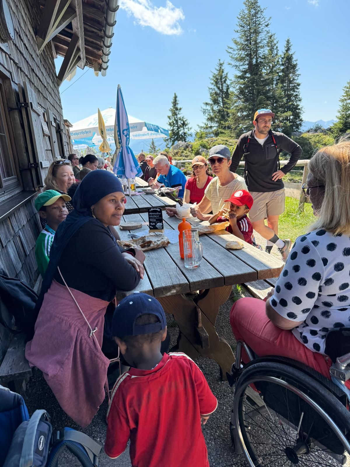 Eine Gruppe von Personen sitzt an einem Holztisch im Freien, umgeben von Bäumen und Bergen. Einige essen, andere unterhalten sich. Ein Kind in rotem Shirt steht am Tisch.