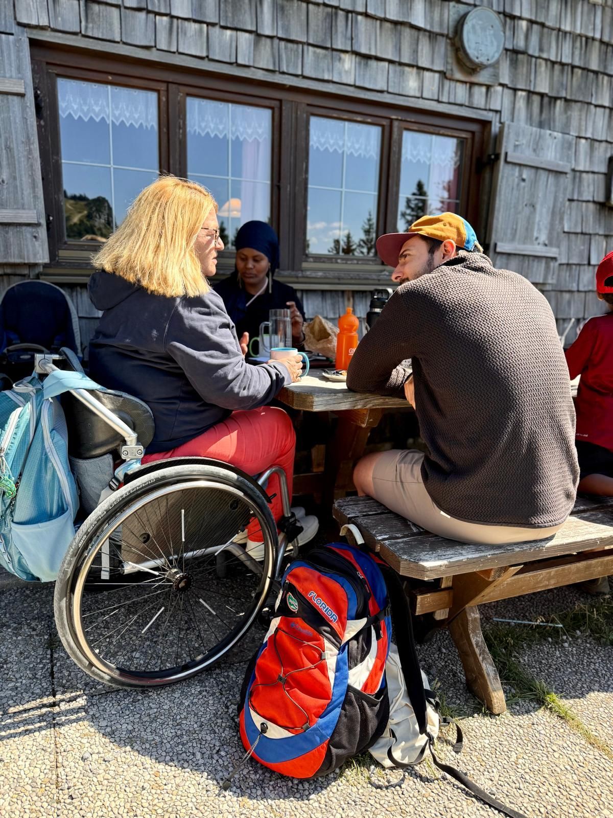 Eine Person im Rollstuhl mit blonden Haaren sitzt an einem Tisch vor einer Hütte. Eine weitere Person in einem braunen Pullover sitzt gegenüber. Auf dem Tisch stehen Getränke und eine orangefarbene Flasche.