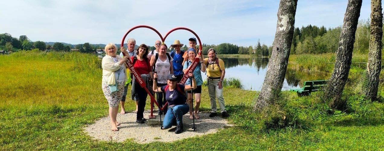 Eine Gruppe von Menschen steht in der Natur. Rechts im Bild fließt ein Fluss. Sie stehen vor einem großen Herz und lächeln. Sie sind auf einer Wanderung unterwegs.