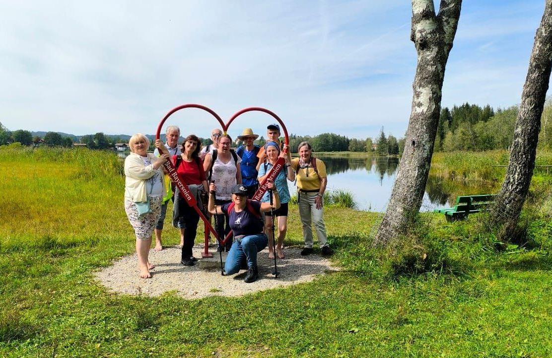 Es ist eine Gruppe Menschen hinter einem Herz aus Metall zu sehen. Sie tragen Wandersachen und stehen auf einer grünen Wiese. Rechts im Bild sind zwei Bäume zu sehen. Im Hintergrund des Bildes läuft ein Fluss.