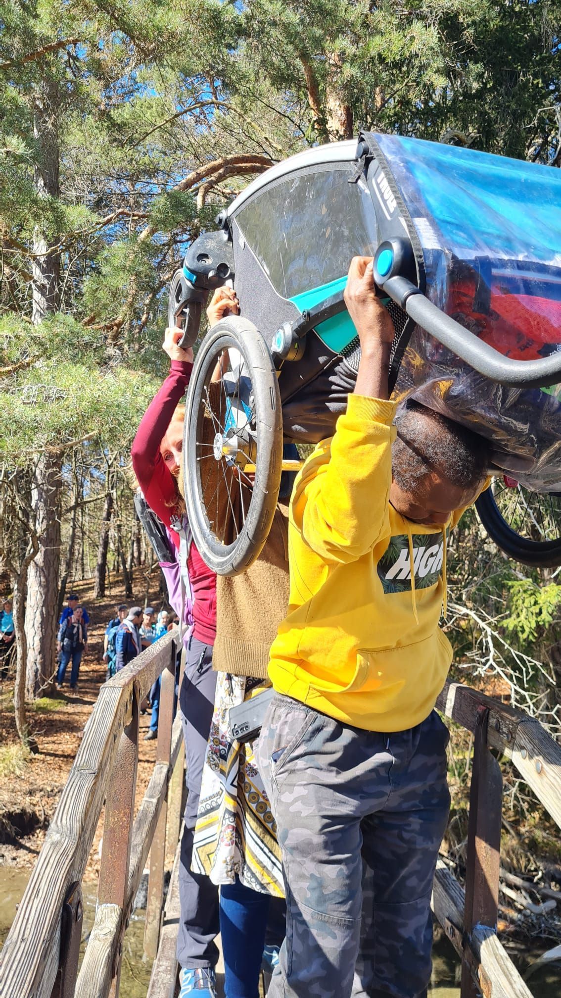 Person in gelbem Pullover hebt einen Kinderwagen über ein Geländer im Wald, weitere Personen helfen dabei