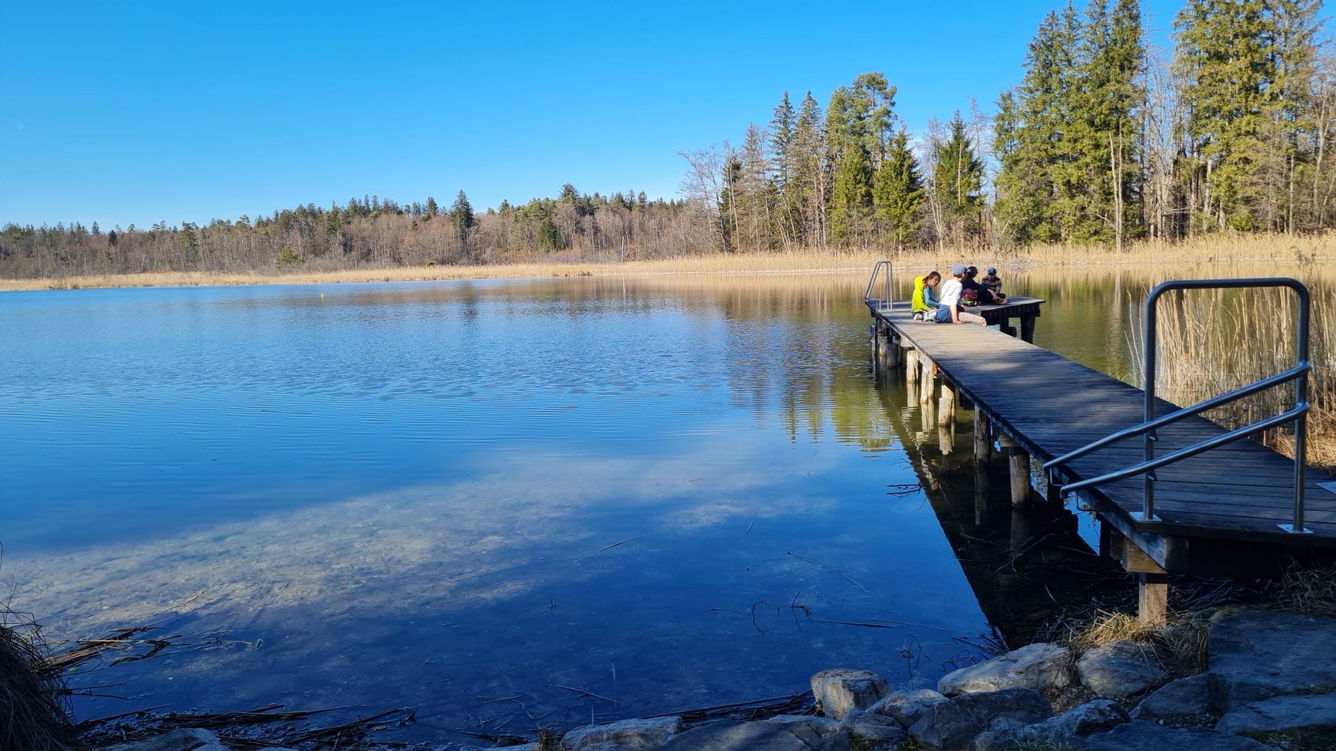 Steg an einem ruhigen See mit drei Personen, umgeben von Bäumen und klarem Himmel