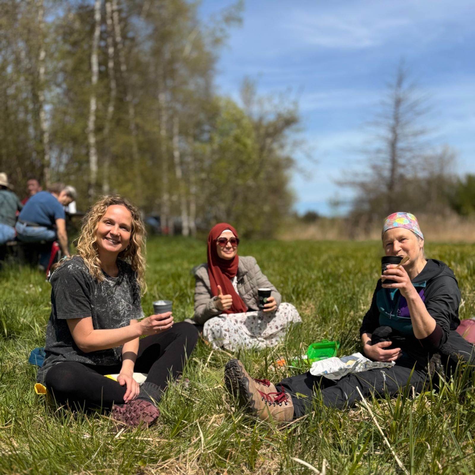 Drei Personen sitzen im Gras, halten Becher in den Händen. Eine Person trägt ein schwarzes T-Shirt, eine andere ein rotes Kopftuch und die dritte eine bunte Kopfbedeckung. Im Hintergrund sind Bäume und weitere Personen sichtbar.