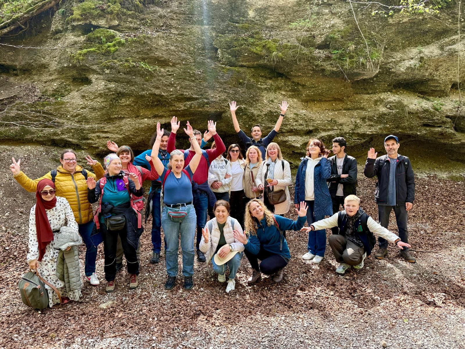 Gruppe von 17 Personen in einem Waldgebiet, einige stehen, andere knien, alle mit erhobenen Händen. Laub und Felsen im Hintergrund.