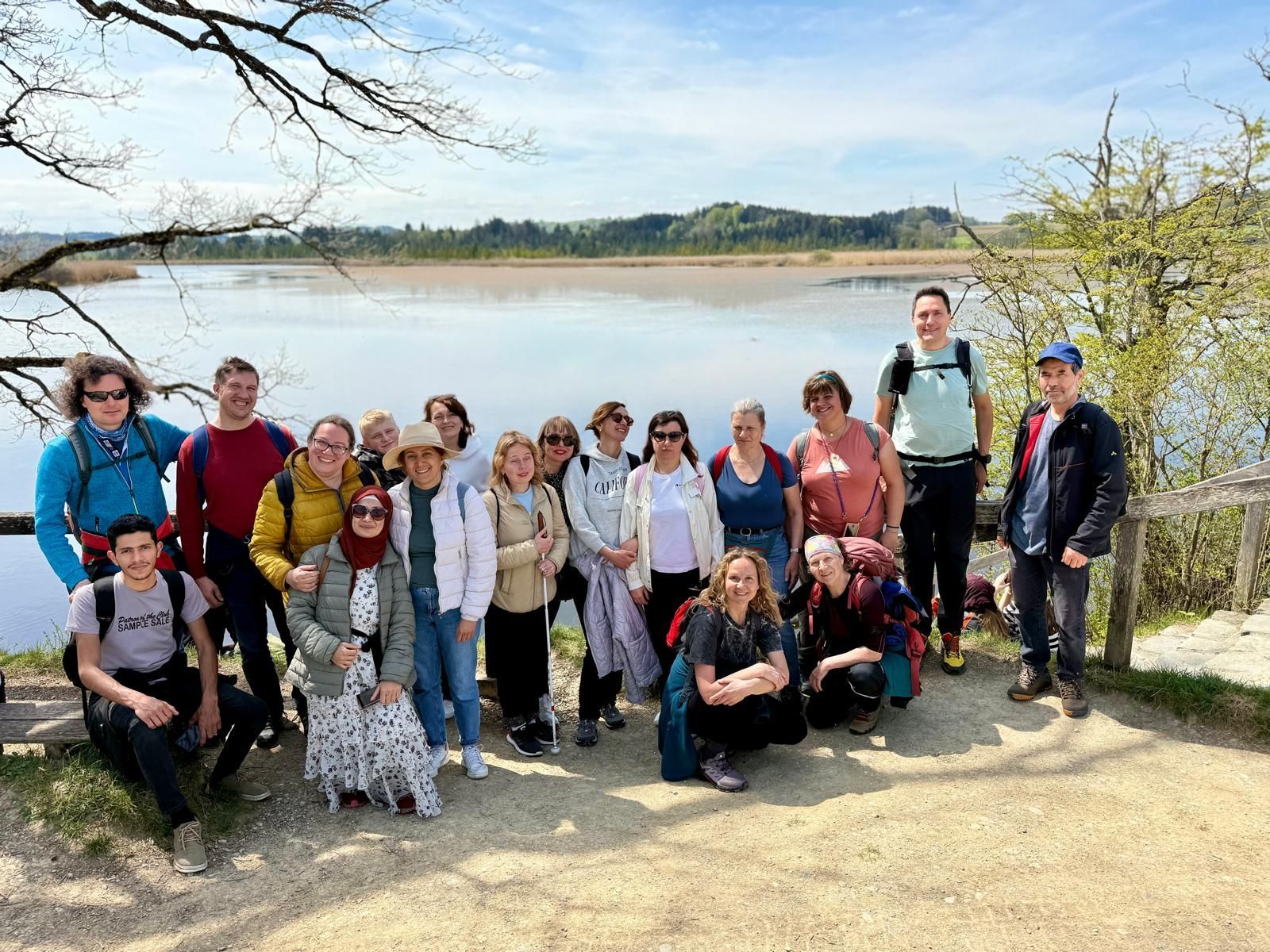 Gruppe von 17 Personen, die auf einem Weg stehen, umgeben von Natur. Im Hintergrund ein Gewässer und Bäume. Einige tragen Rucksäcke und Hüte.