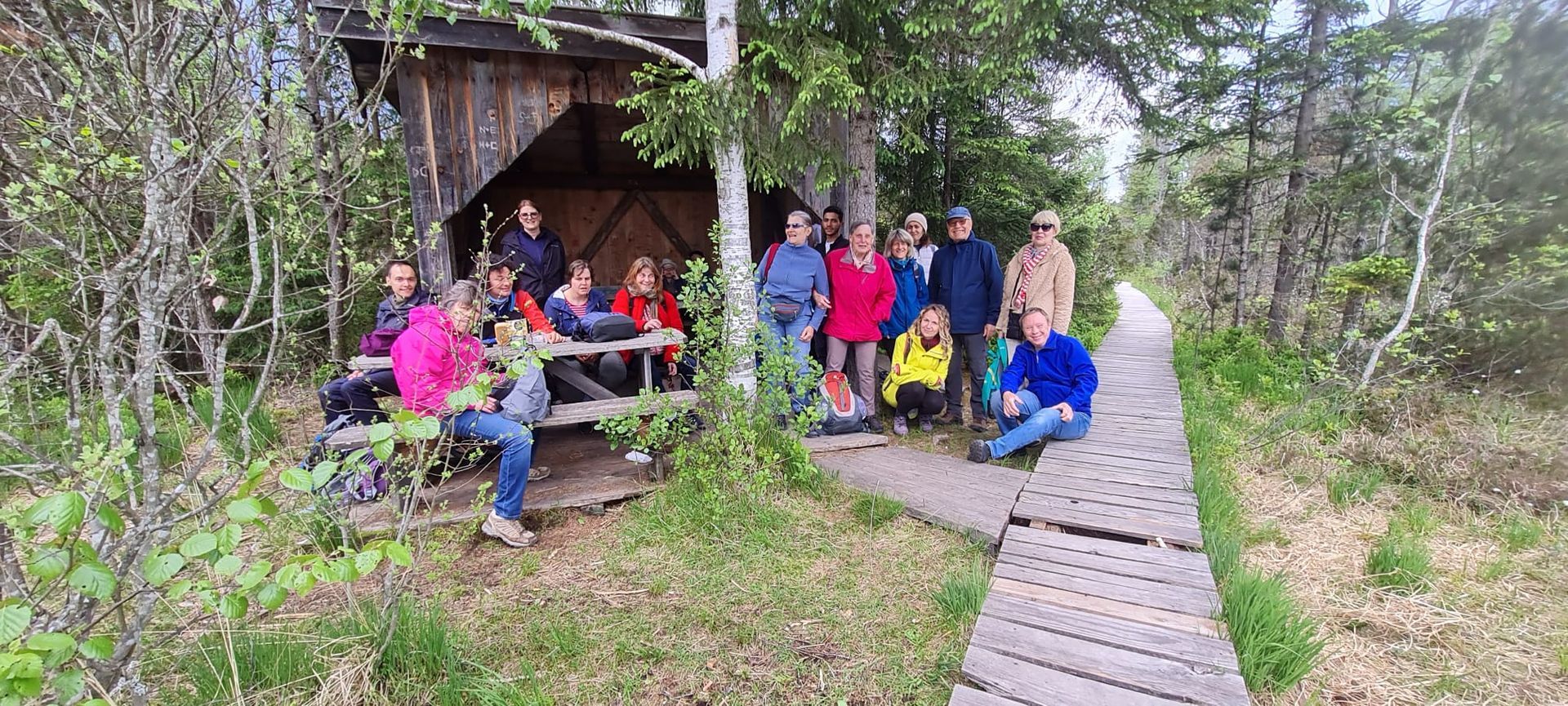 Gruppe von 13 Personen sitzt an einer Holzbank unter einem überdachten Pavillon im Wald. Ein Holzsteg führt in den Hintergrund.